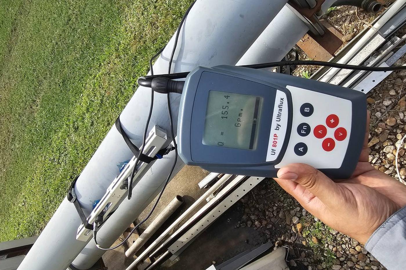 Ultraflux UF801-P portable clamp-on flow meter displaying Q = 155.4 GPM with rail-mounted transducers on the pipe behind