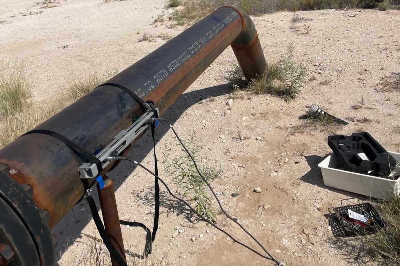 Clamp-on transducer rail strapped to a corroded carbon steel pipe in the West Texas desert, instrument case open on the ground